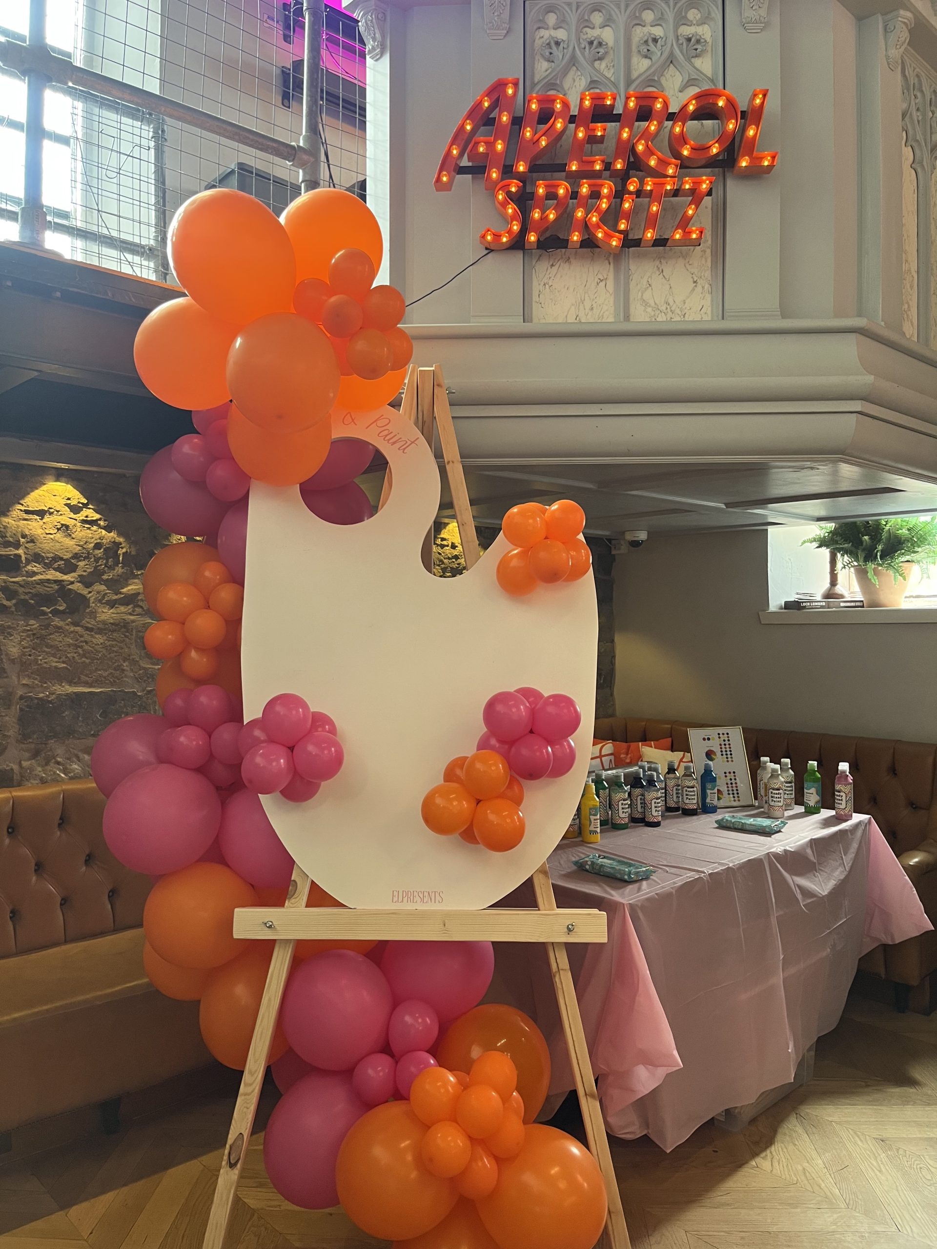 Large easel sign with orange and pink balloons in front of an Aperol sign